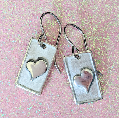 ONE HEART EARRINGS | <Z-FIRE.COM> A polished and highlighted silver heart on frosted silver rectangle background with polished edges. .950 Fine Silver Rectangles About 3/8" Wide About 9 mm Wide Approximately 5/8" Total Length About 1.6 cm Total Length .925 Sterling Silver Ear Wires
